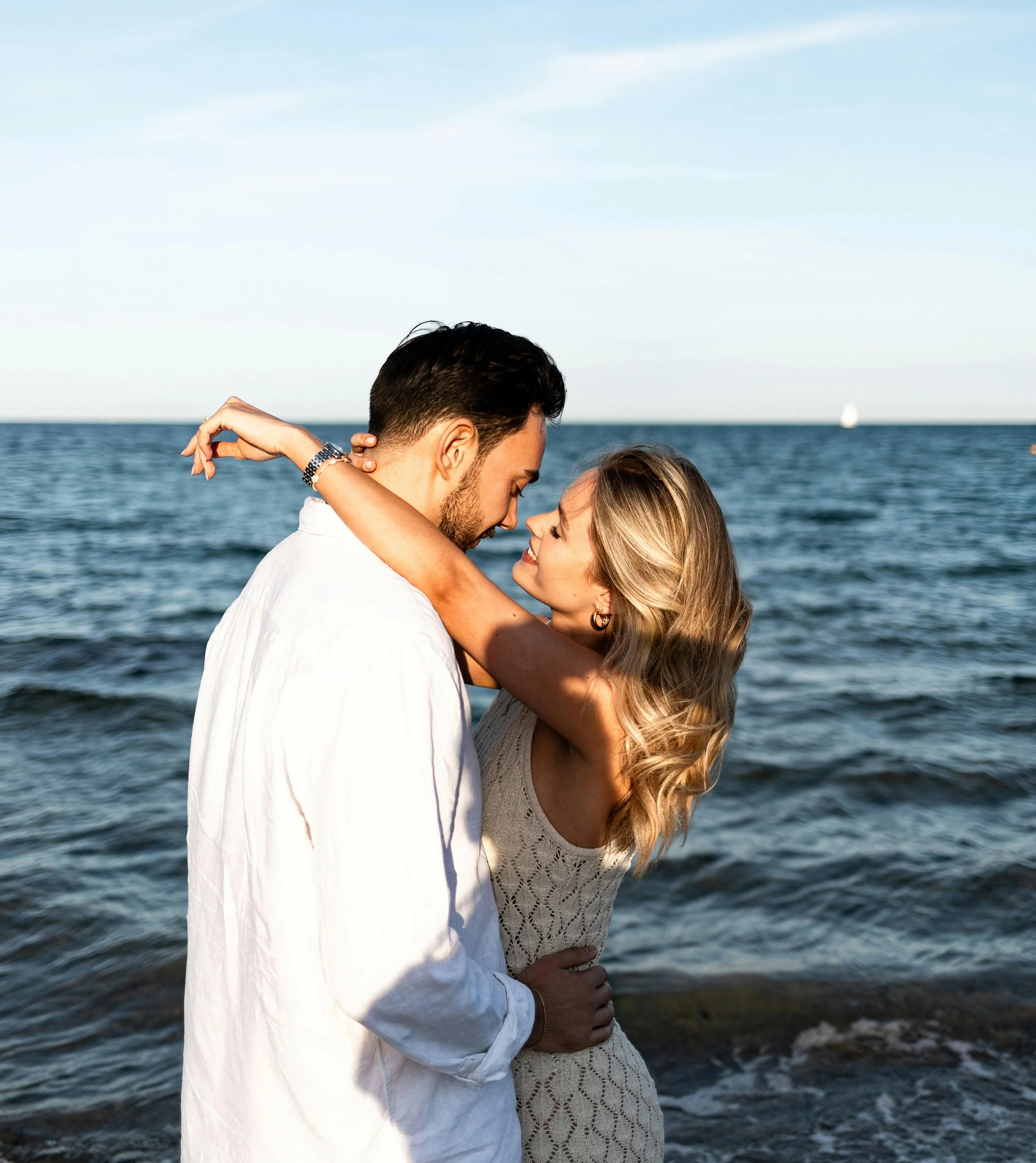A couple embracing on the beach during sunset, standing close to the water with the ocean in the background.
