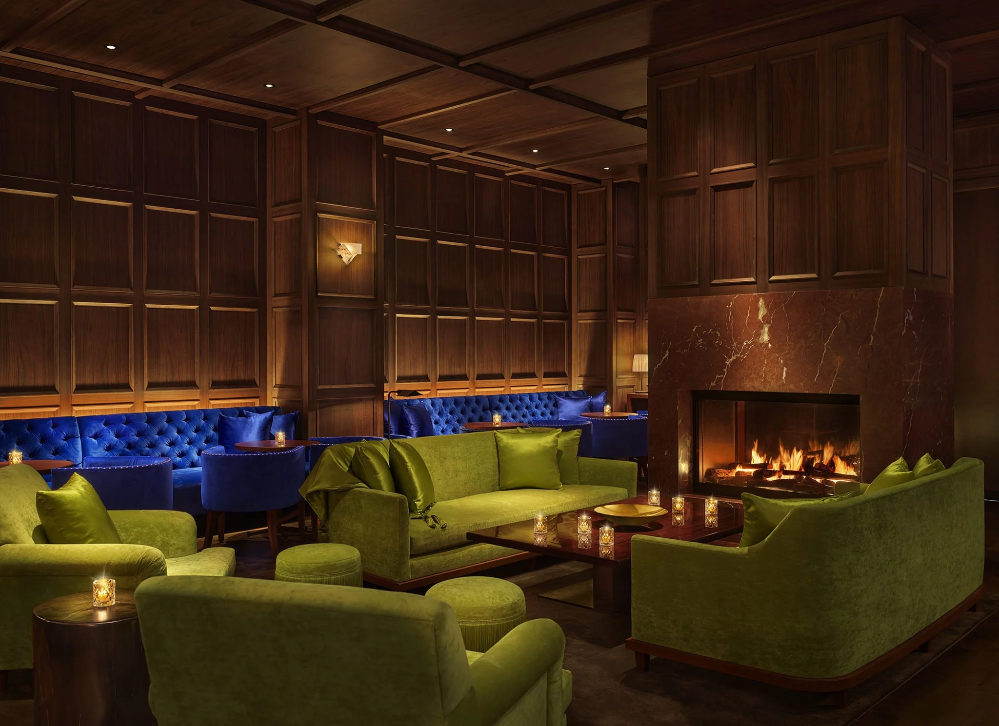 A luxury lounge with green sofas, a fireplace, and blue velvet chairs along the wall in a wood-paneled room.