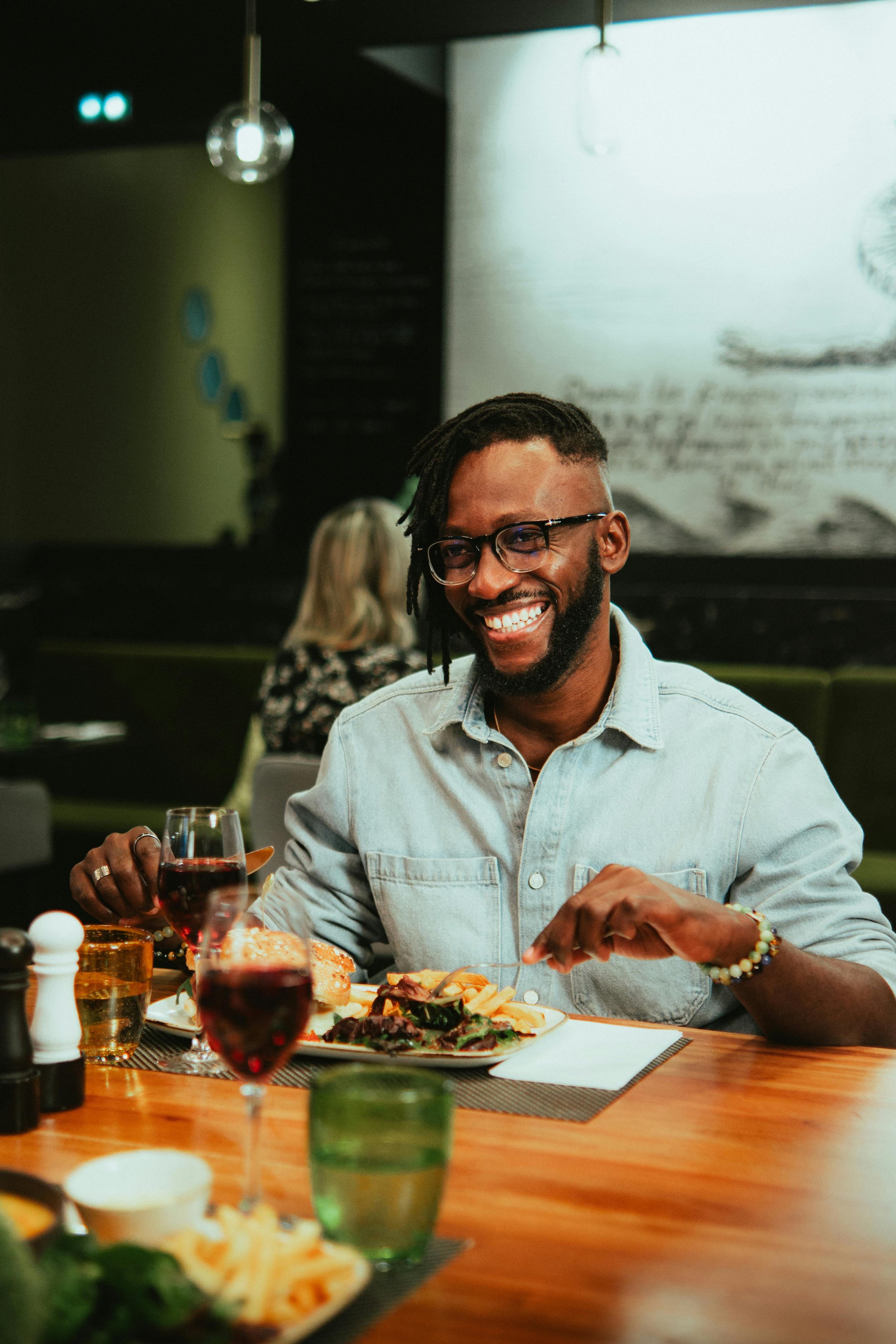 A man with glasses, a beard, and dreadlocks smiling while enjoying a meal at a restaurant, with a glass of red wine, a plate of food, and other drinks and dishes on the table.