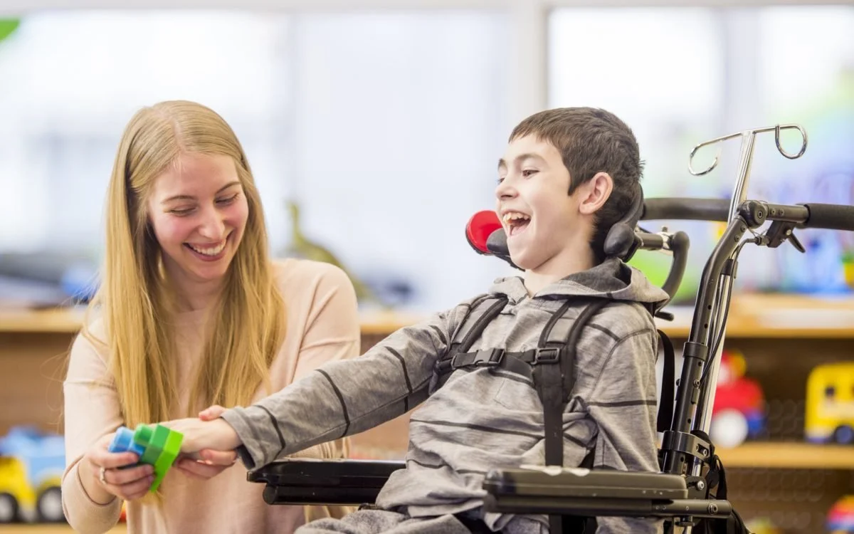 A young boy in a wheelchair with a medical headrest, smiling and holding colorful stacking cups, with a smiling woman assisting him in a room filled with toys and colorful objects.