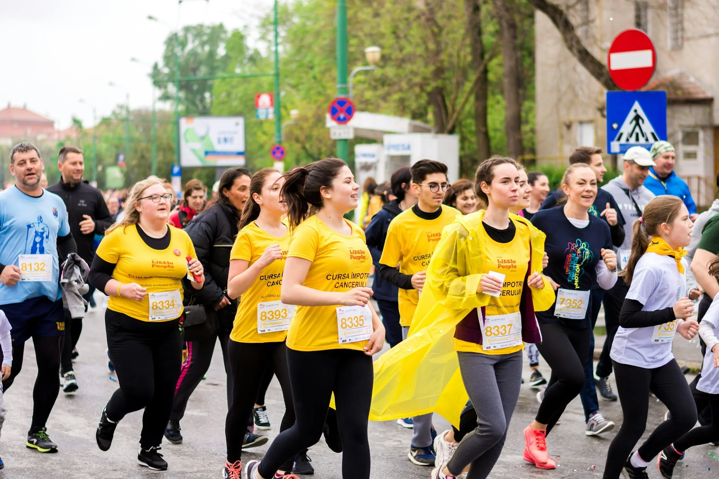 A group of runners participate in a marathon, wearing brightly colored athletic shirts and bib numbers, with some wearing yellow shirts supporting cancer awareness.