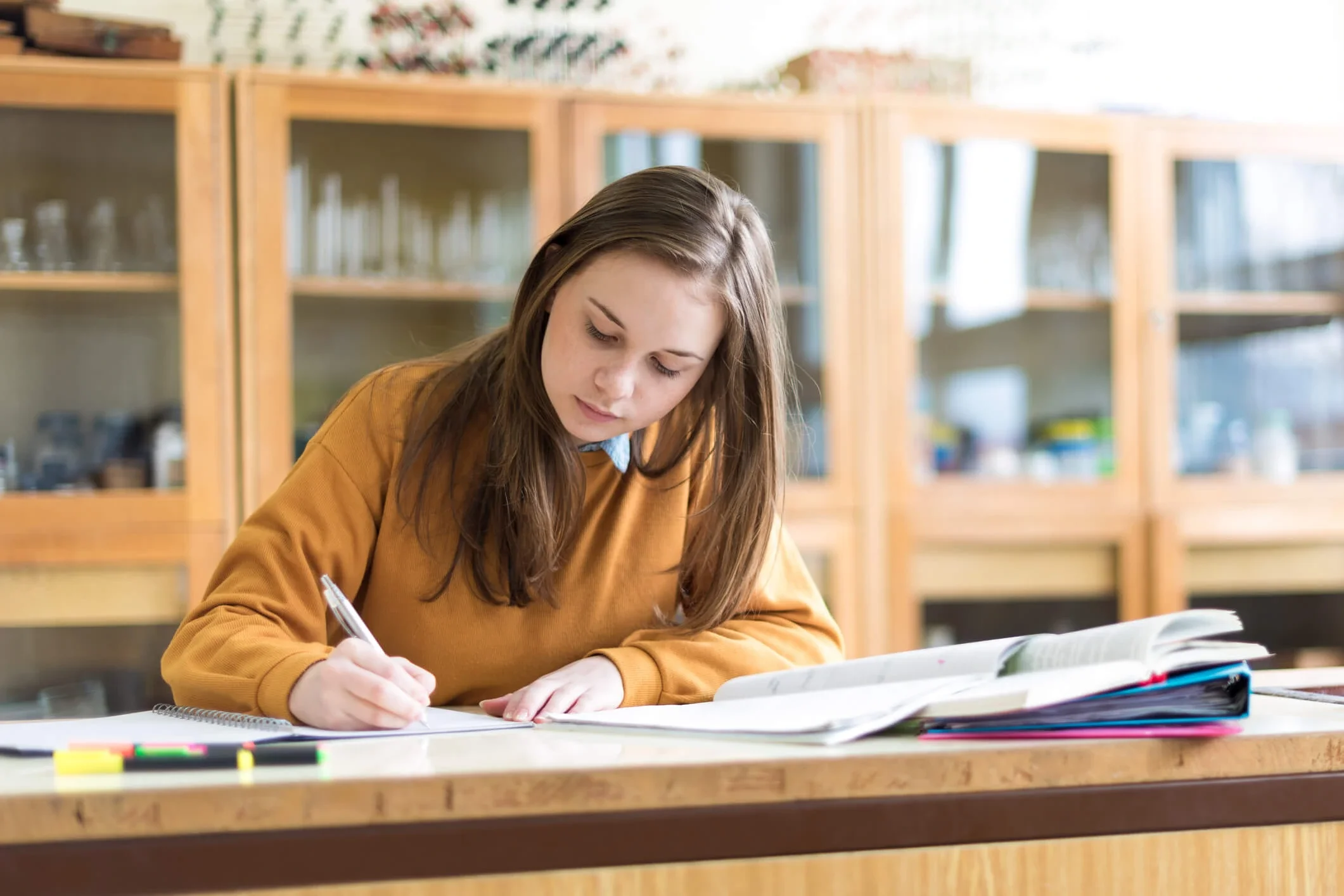 A young woman with long brown hair, wearing a mustard yellow sweater, is studying and taking notes at a table in a library or classroom. There are books and colored markers in front of her, with wooden shelves filled with glass display cases in the background.