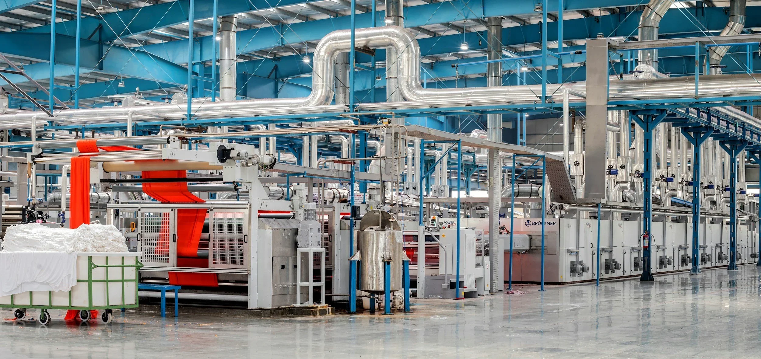 Industrial manufacturing facility with blue steel framework and large silver pipes, machinery, and a conveyor belt system processing fabric or similar material.
