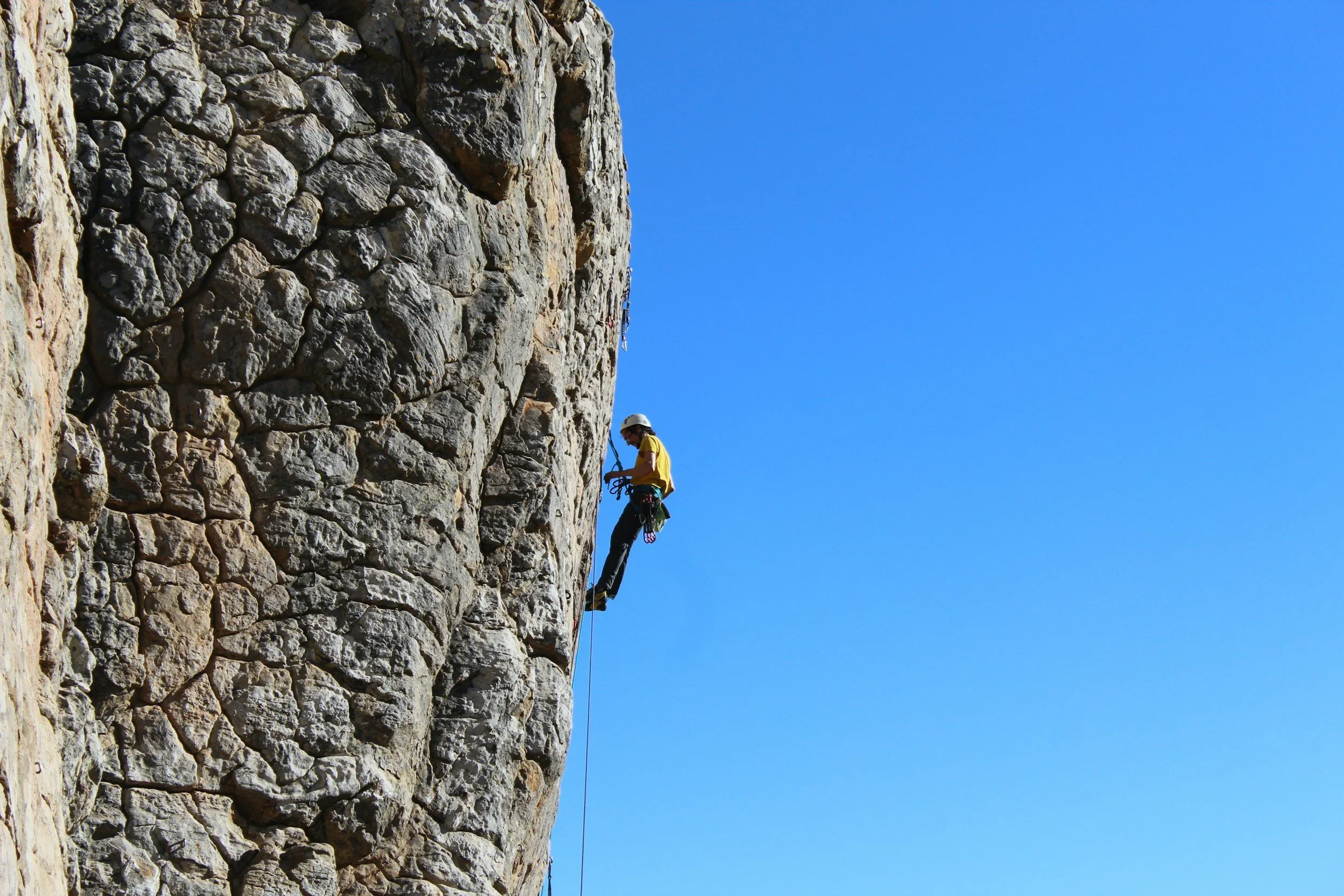 A person wearing a helmet and climbing gear is rappelling down a large, rocky stone wall on a bright, sunny day with a clear blue sky.