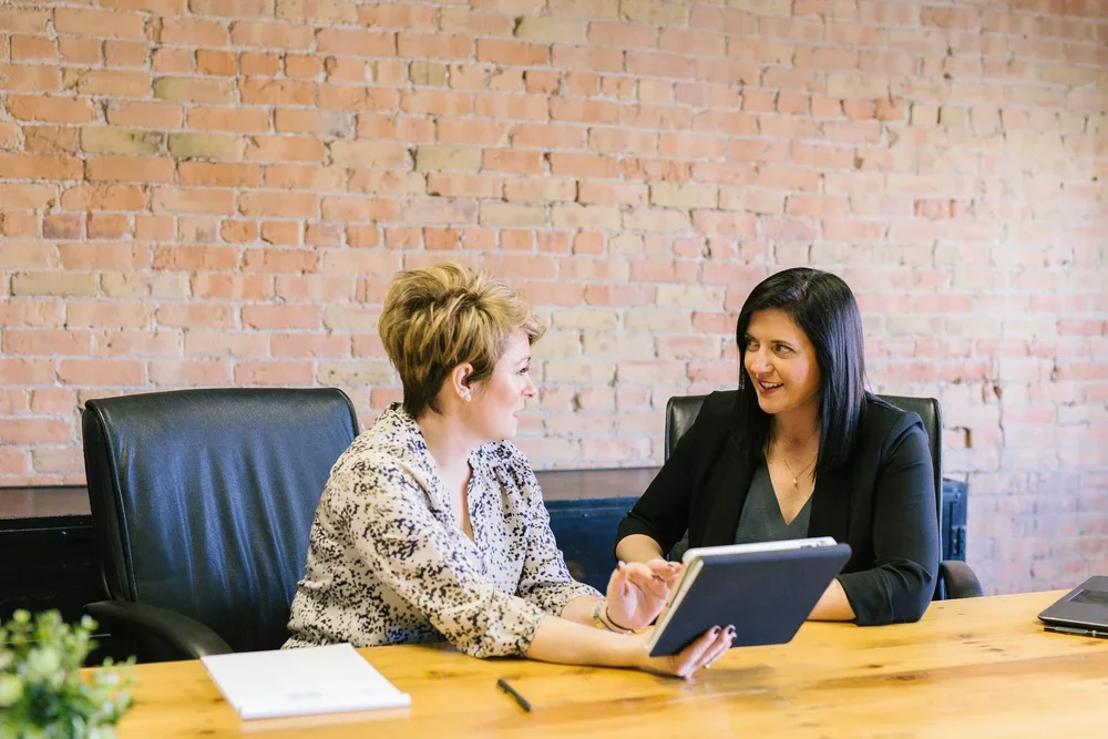Two women sitting at a wooden table having a discussion, one holding a tablet, against a brick wall background.
