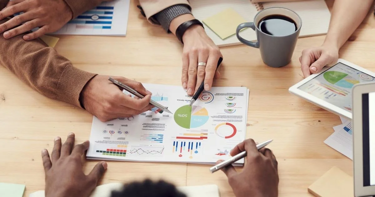People analyzing financial reports and data charts at a wooden table with tablets and a coffee mug.