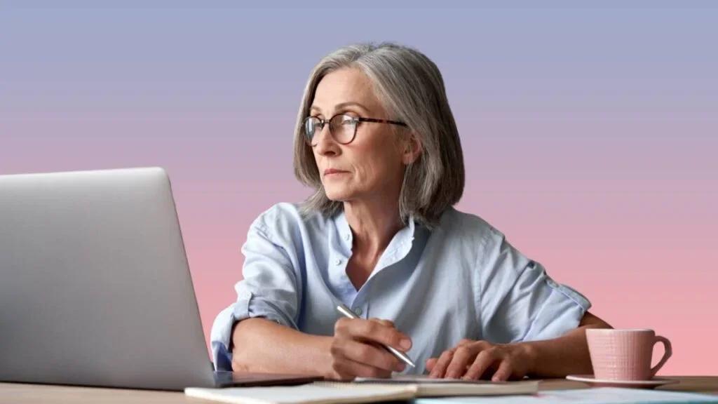 An older woman with gray hair and glasses working on a laptop at a desk, writing notes in a notebook, with a pink mug nearby, against a blurred purple and pink gradient background.