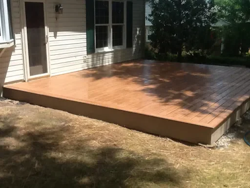 Wooden deck attached to the back of a house with sliding glass door and windows, surrounded by a yard with grass and trees.