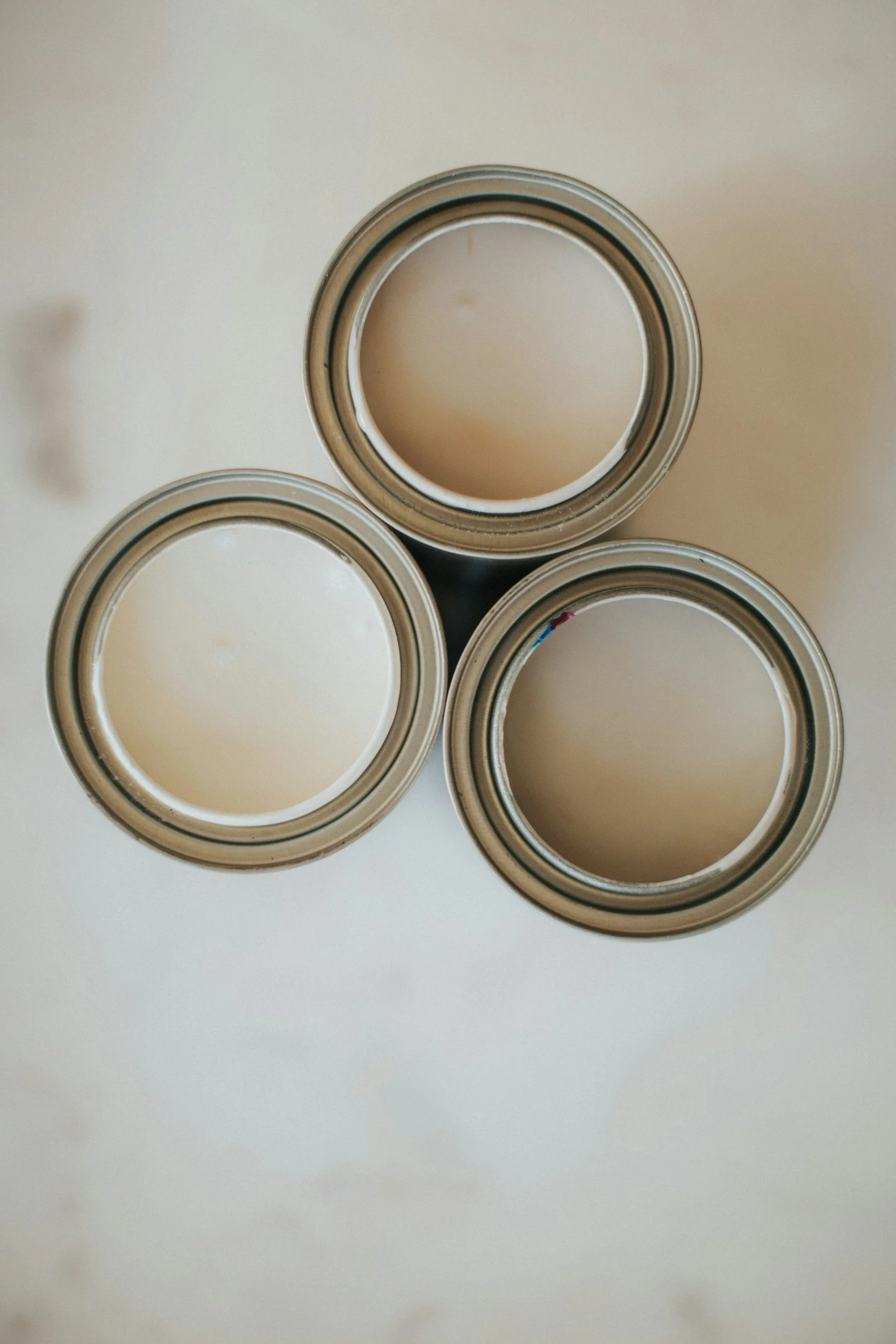 Top view of three paint cans filled with beige paint arranged in a triangle on a white surface.