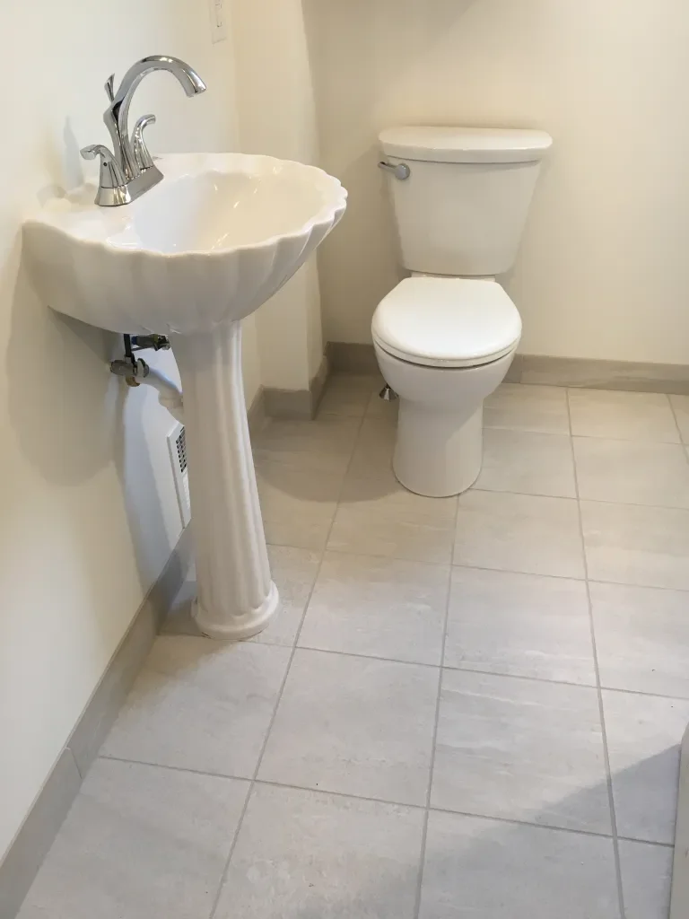 A small bathroom with a pedestal sink and a toilet, both white, on a tiled floor with light-colored tiles.