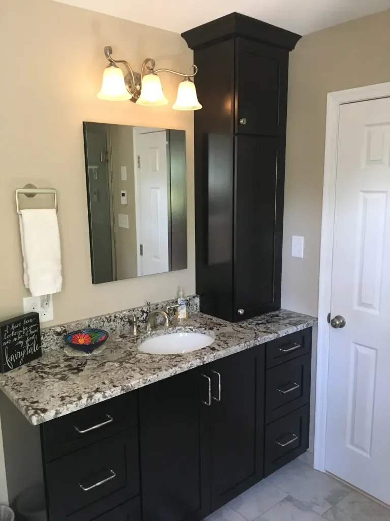 Bathroom vanity with a granite countertop, black cabinets, a mirror, a faucet, three drawers, a decorative bowl, a small sign, a towel, and a wall-mounted light fixture.