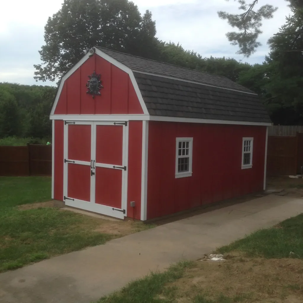 Red and white miniature barn with black decorative emblem on front, two small windows, and a sloped shingled roof, situated on a lawn next to a concrete pathway.
