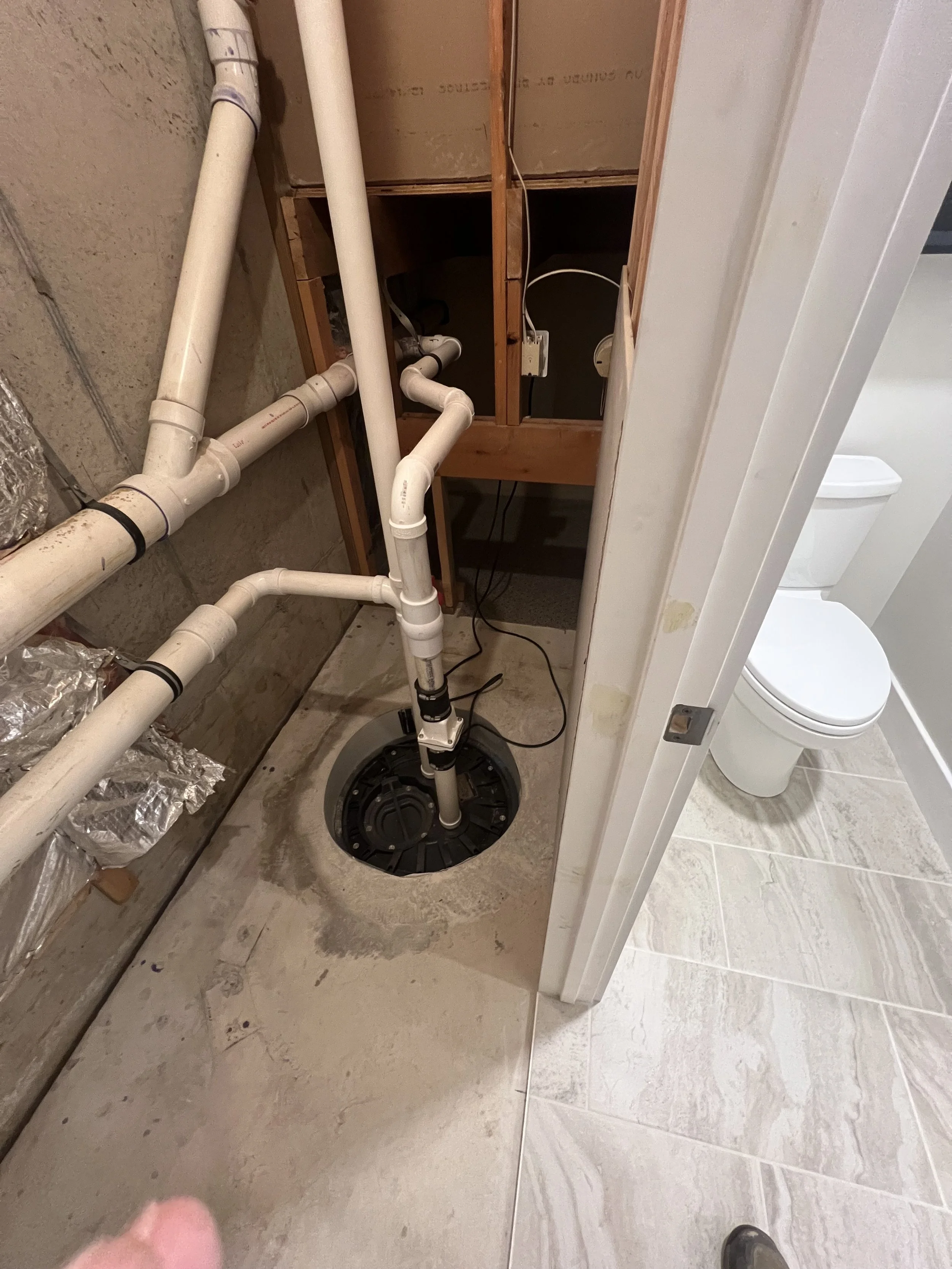 Plumbing pipes and sump pump installation in a bathroom utility closet with a view of a toilet in the adjacent bathroom.