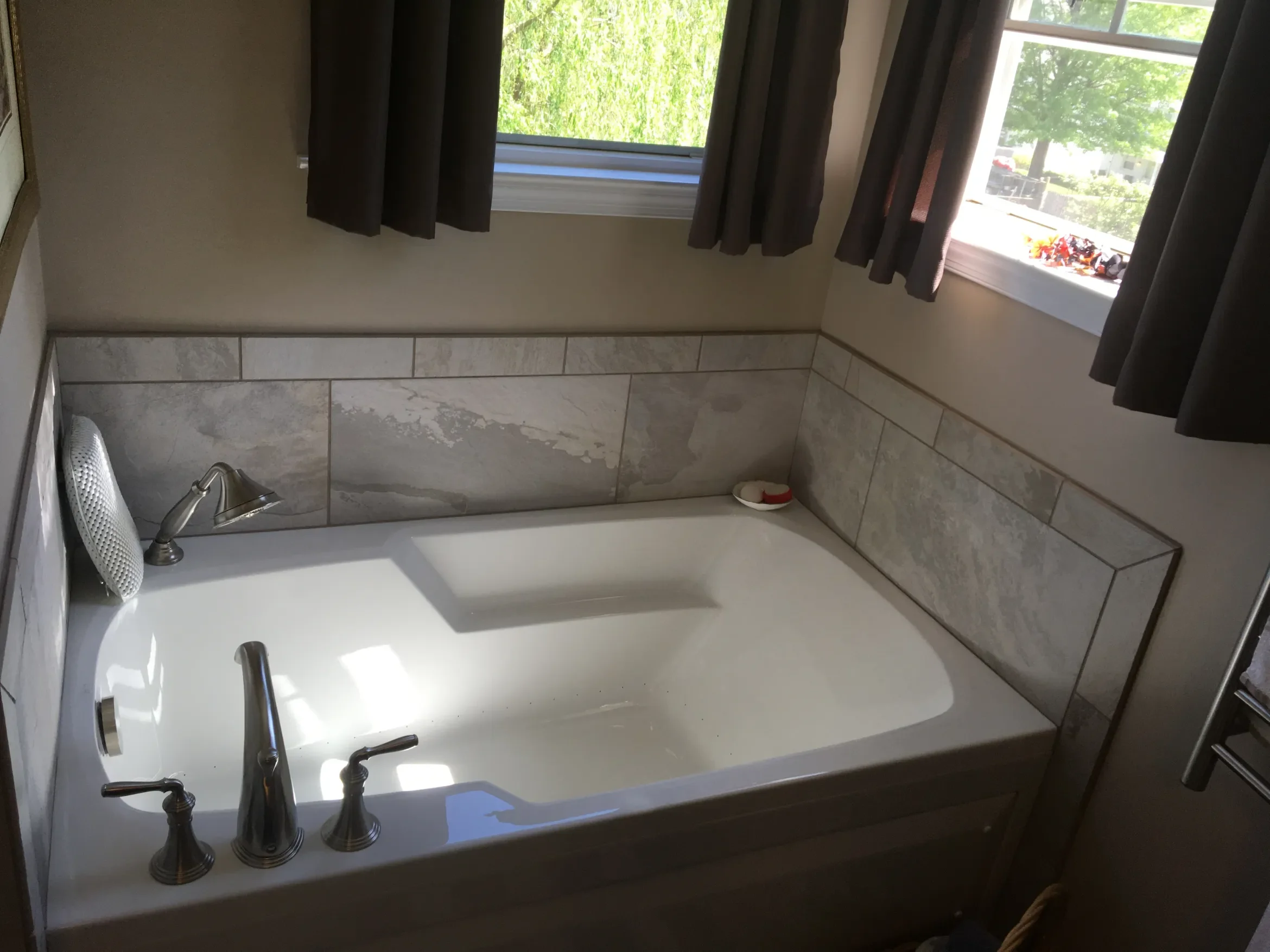 A bathroom with a white bathtub, silver fixtures, beige walls, and a large window with dark curtains. Outdoors, green trees and a parking lot are visible.