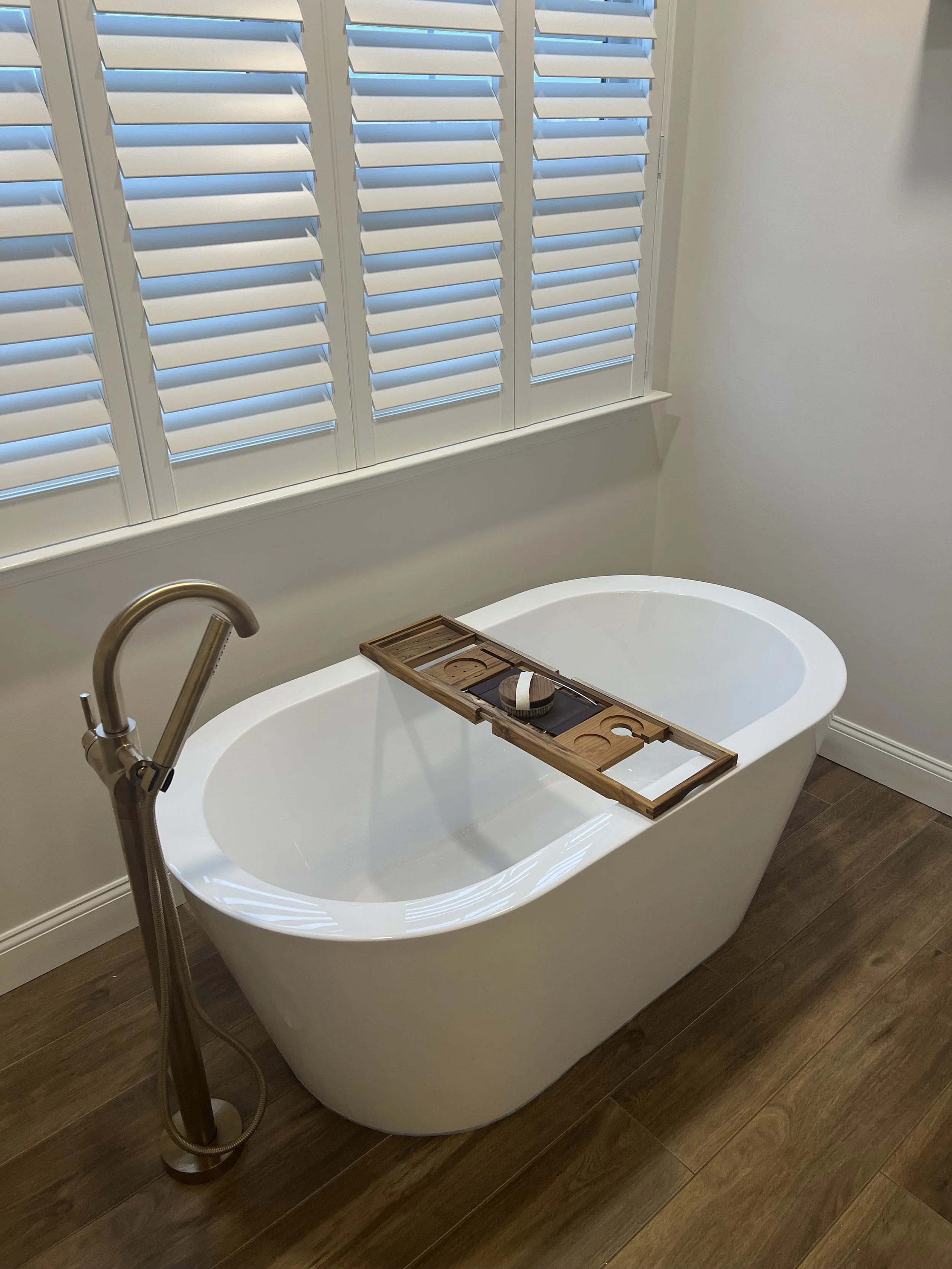 Bathtub with a wooden bath tray holding a soap and a sponge, next to a window with white blinds, in a bathroom with wood flooring.