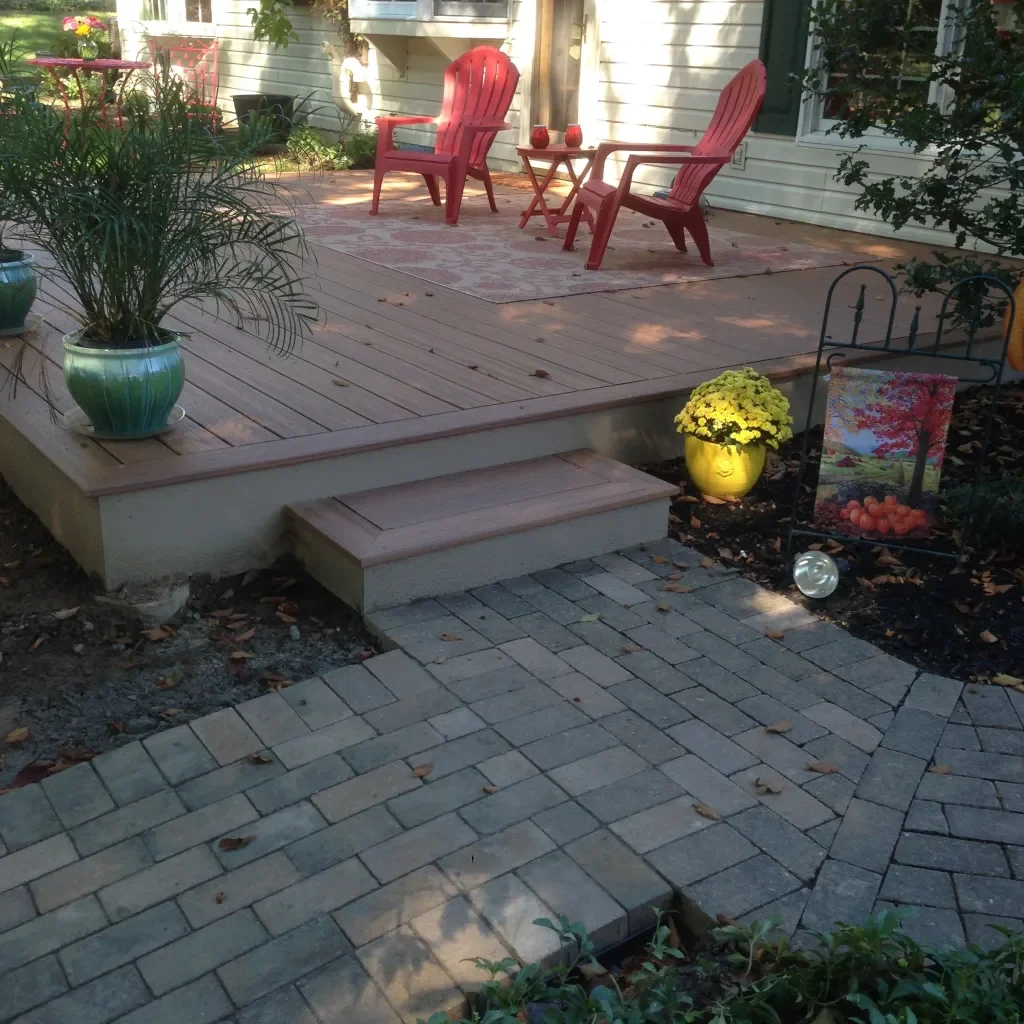A backyard patio scene with a raised wooden deck, two red Adirondack chairs, a small side table with candles, and potted plants. There are brick pavers leading to the deck and decorative items like a yellow flower pot and a garden flag.