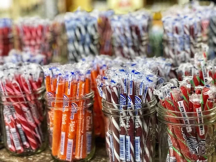 Glass jars filled with various striped candy sticks in red, orange, blue, white, and green colors, displayed on a store shelf.