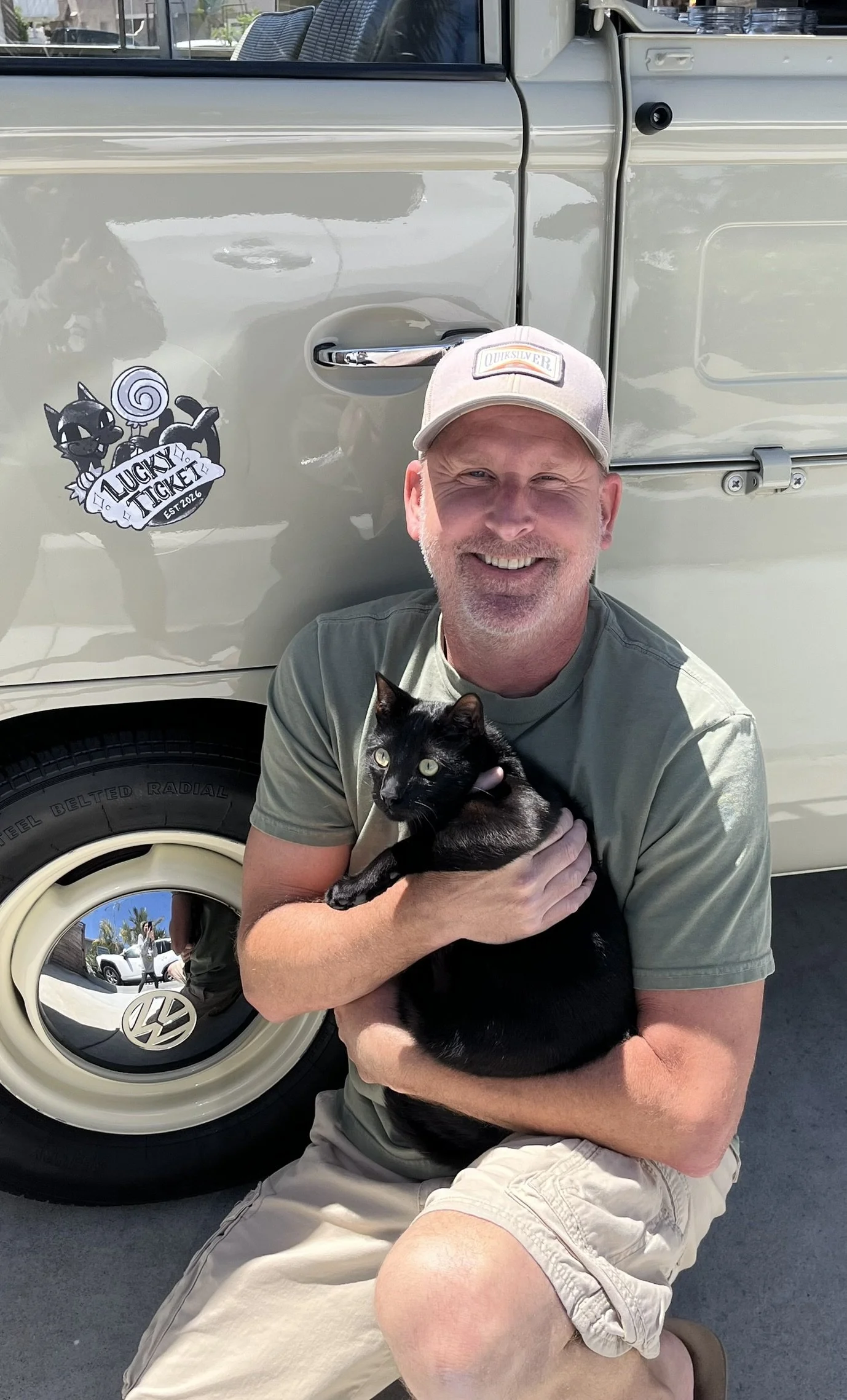 A man smiling while holding a black cat with green eyes in front of a vintage beige Volkswagen vehicle. The man is wearing a light-colored Quiksilver cap and a gray T-shirt. The vehicle, which features a cartoon sticker of a black cat and a lollipop with the text "Lucky Ticket Est. 2016," is parked outside, with visible reflections of the surrounding area in the car's shiny surface.