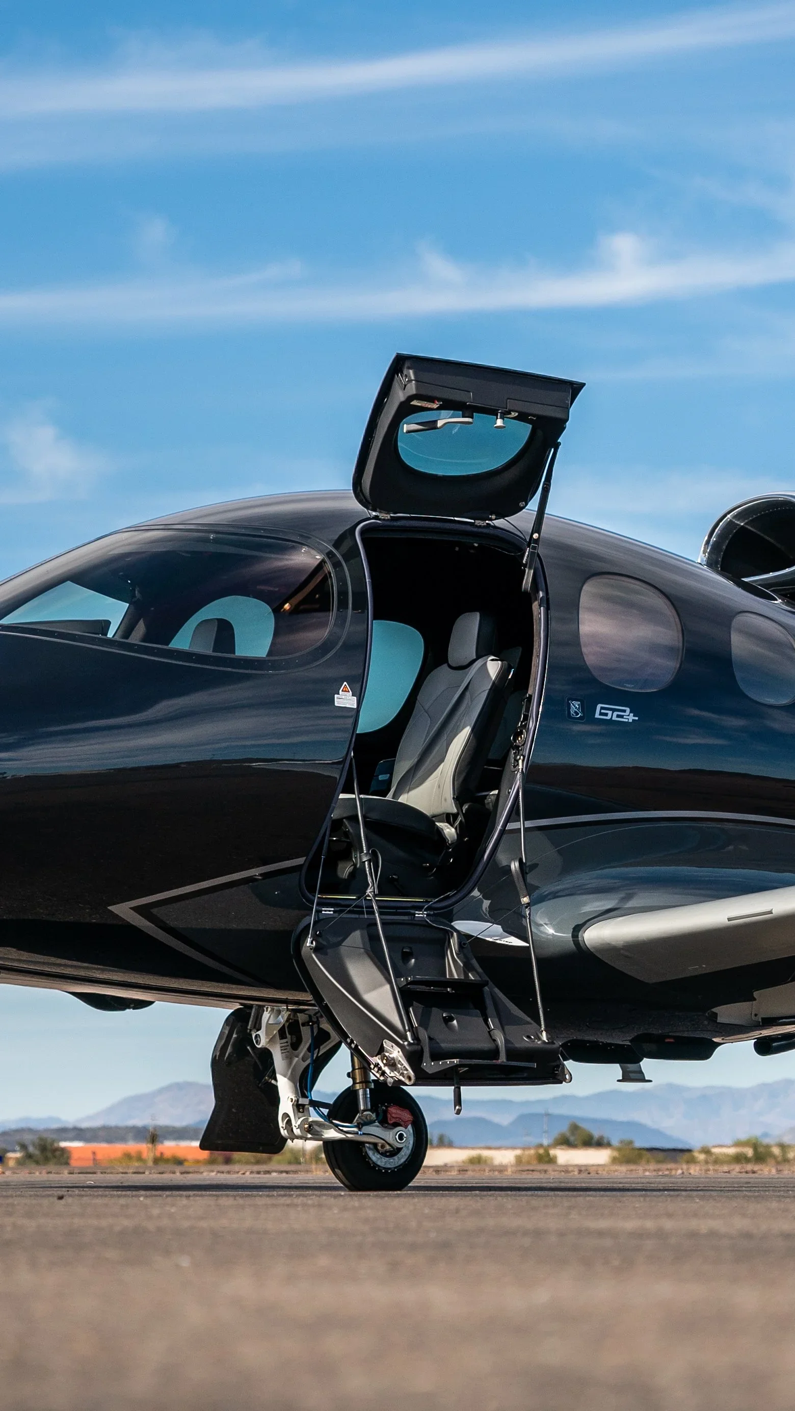Close-up of a black private jet aircraft with the cockpit door open, showing the interior seat and the open stairs, parked on a tarmac with mountains and a blue sky in the background.