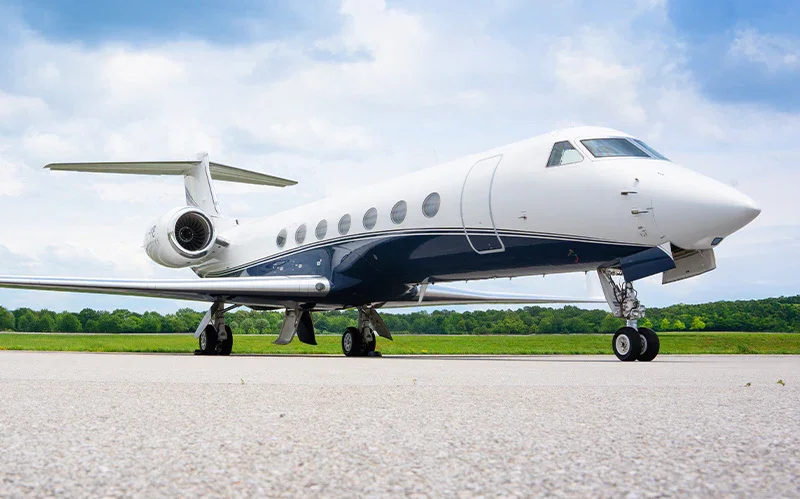 A white and blue private jet on a runway with greenery and a partly cloudy sky in the background.