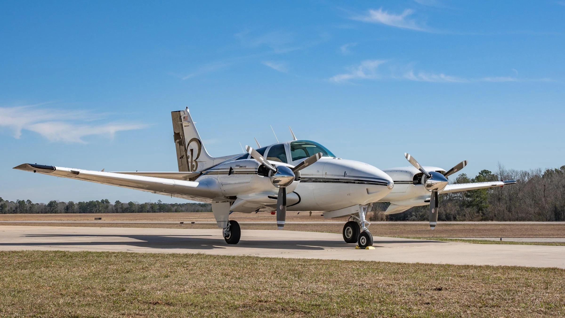 A silver twin-engine propeller airplane parked on a tarmac with a clear blue sky and trees in the background.