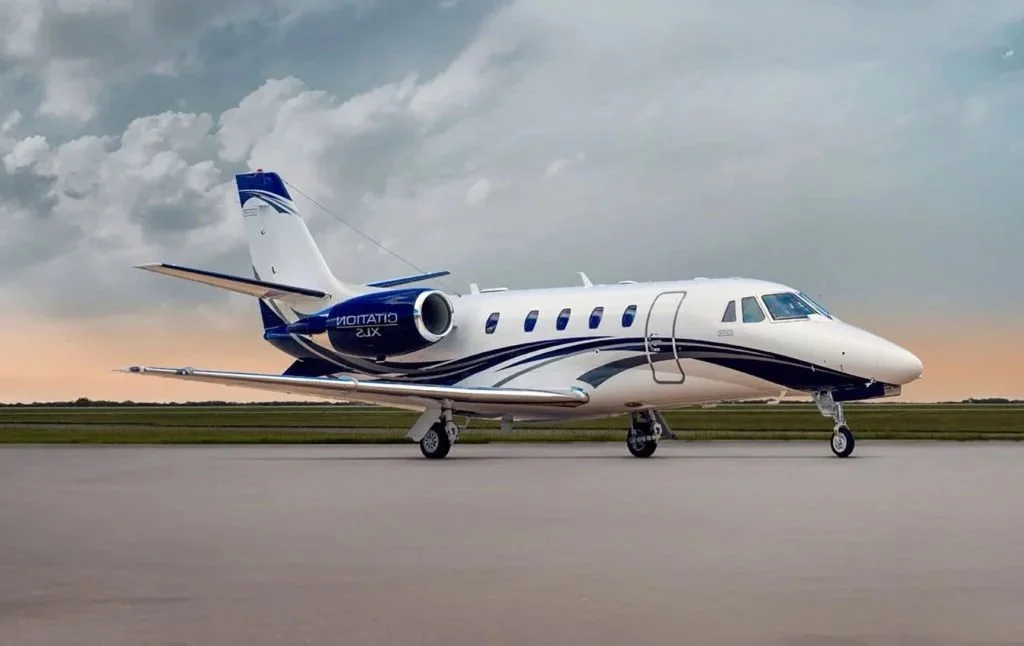 A private jet aircraft parked on an airport taxiway, with a cloudy sky and sunset in the background.