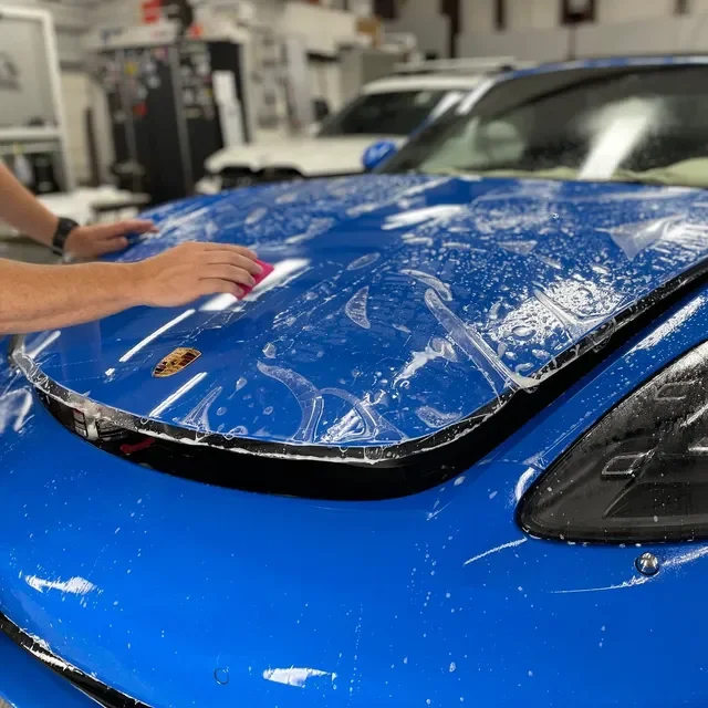A person installing a clear protective film on the hood of a blue Porsche car in an automotive workshop.