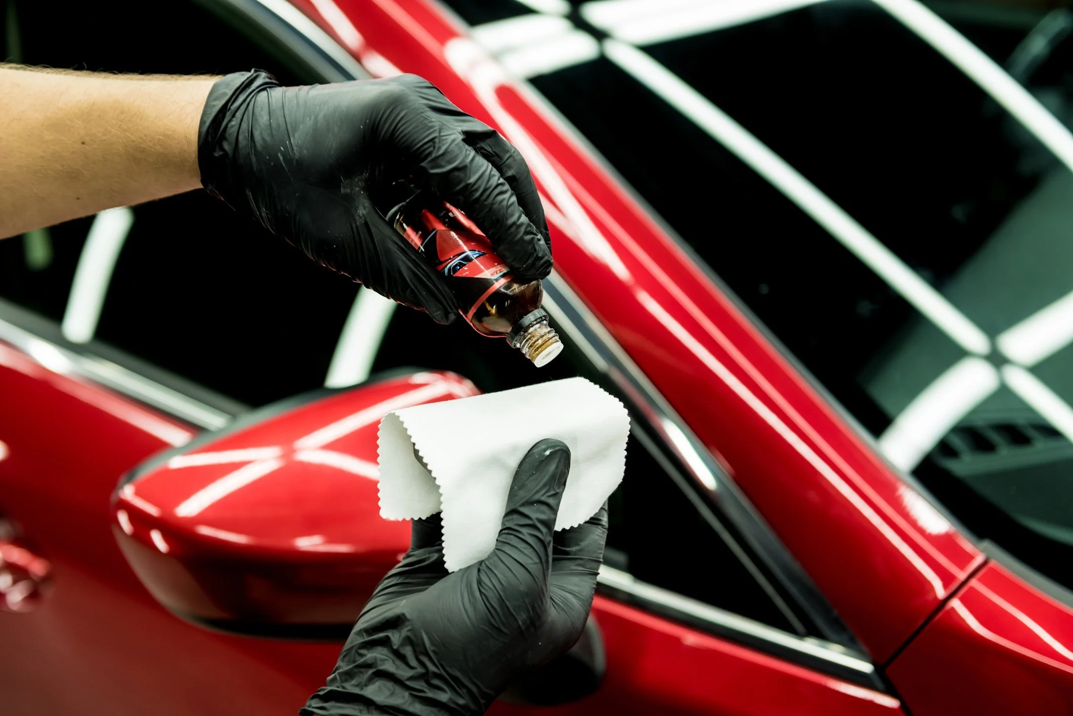 Person wearing black gloves applying a liquid from a small bottle onto a white cloth near a red car's side mirror.