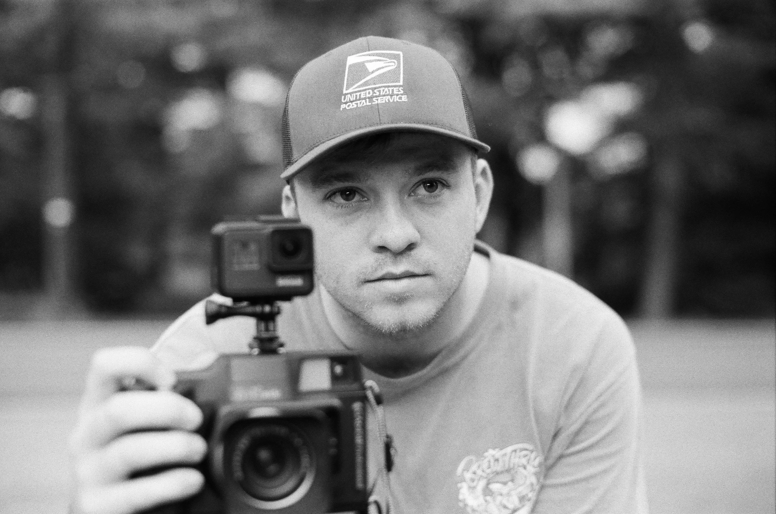 A young man with a serious expression holding a camera, wearing a cap with the United States Postal Service logo, outdoors with blurred trees in the background.