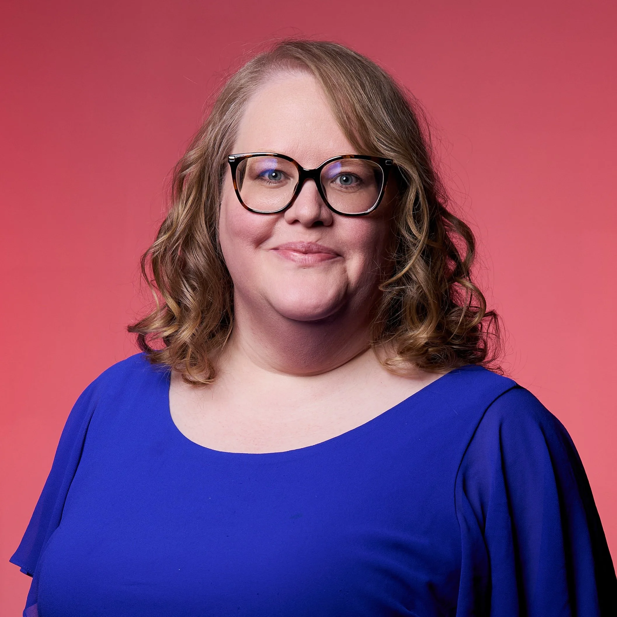 A woman with shoulder-length curly hair, wearing glasses and a royal blue top, posing against a pink background.