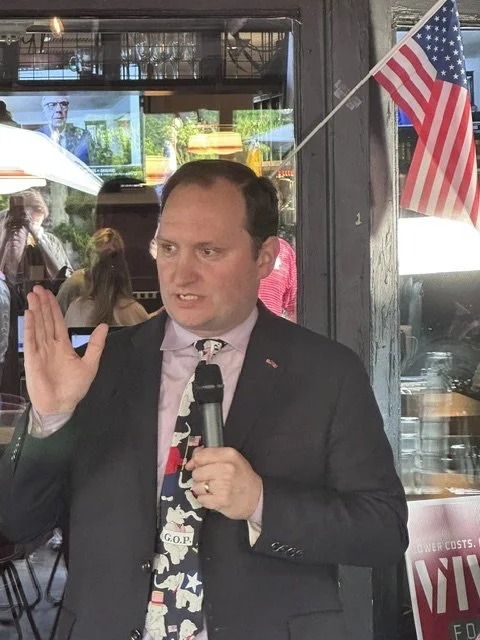 Man in a suit speaking into a microphone with an American flag in the background.