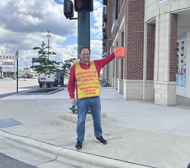 A man standing on the sidewalk holding an orange container, wearing a bright yellow vest that says 'Knights of Columbus Helps' and 'Developmentally Disabled Citizens'. He is standing near a traffic light and a building with red brick and beige stone exterior.