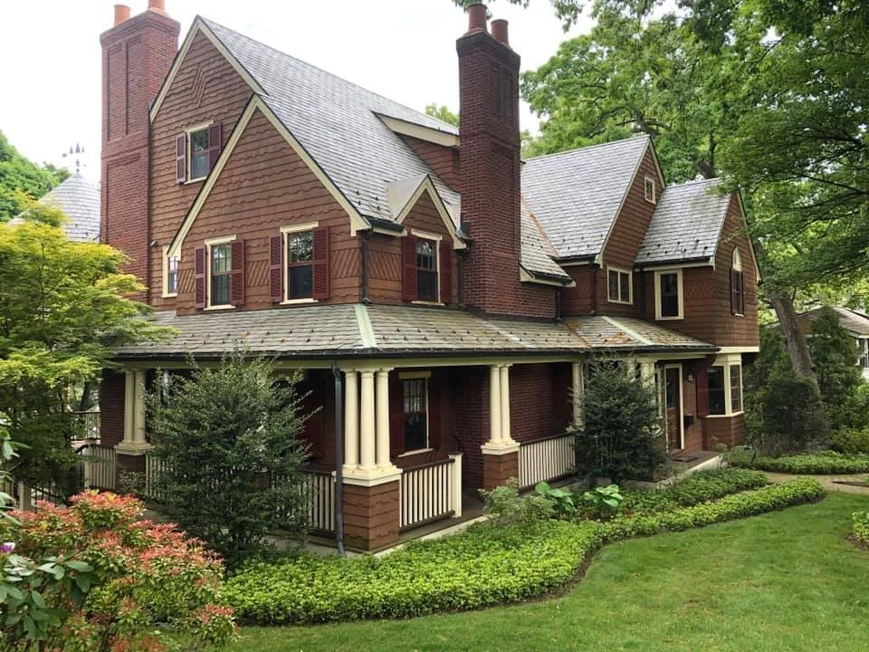 A large two-story house with a brick facade, multiple gabled roofs, and a wraparound porch with white columns, surrounded by green trees and a well-maintained lawn.