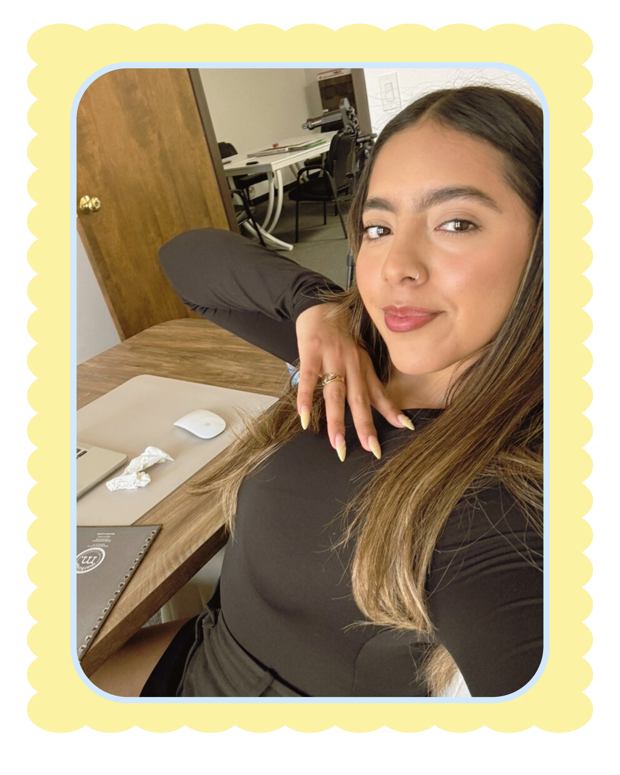 A young woman with long brown hair, wearing a black long-sleeve shirt, sitting at a wooden table in an office or workspace. She is smiling slightly and looking at the camera, with her head resting on her left hand. On the table, there is a white computer mouse, a laptop, and some tissues. The background shows chairs, a doorway, and office furniture.