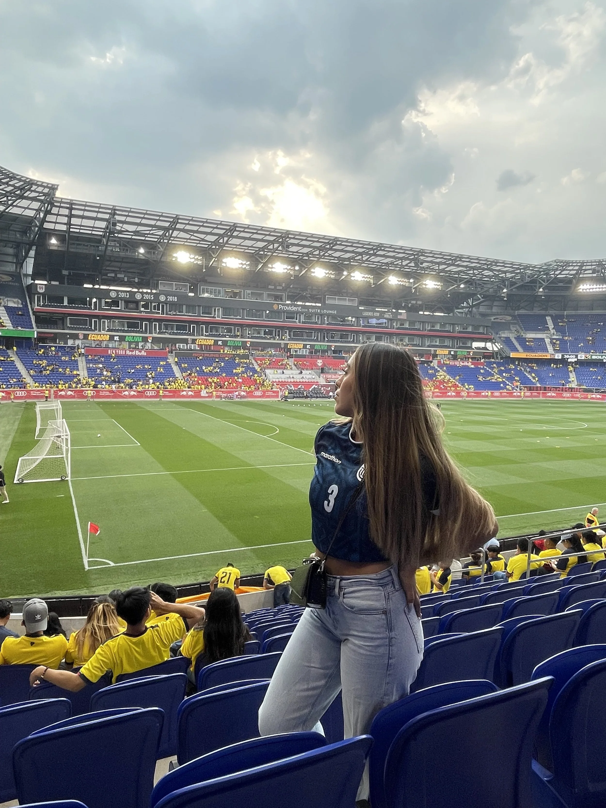 View of a soccer stadium filled with spectators, some wearing yellow jerseys, with a woman standing and looking on from the seats. The field is green with goalposts visible, and the sky above is cloudy.