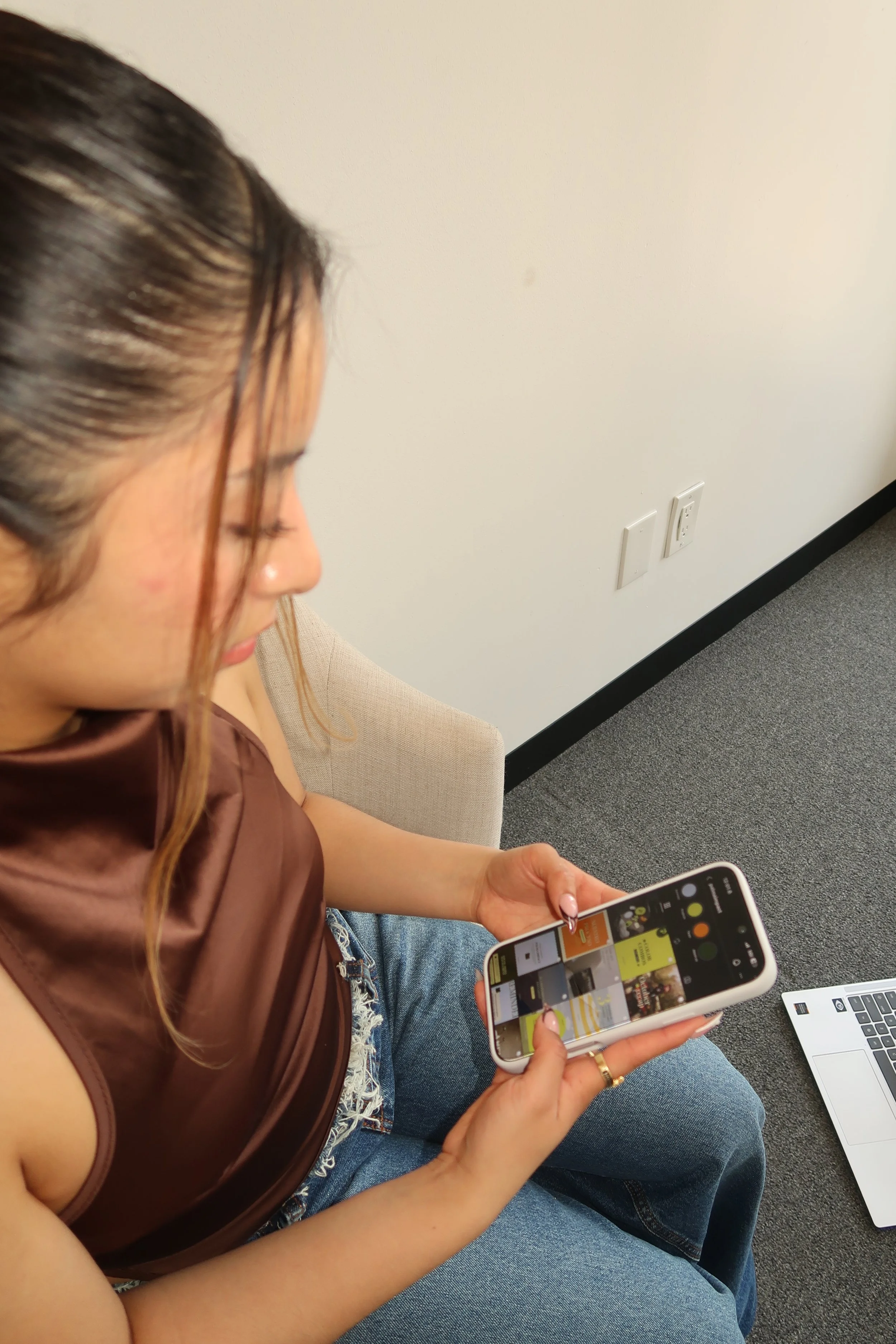 A woman sitting on a chair in an office, looking at her smartphone, with a laptop on her lap. She has long, light brown hair and is wearing a sleeveless brown top and blue jeans. The office wall is plain white with electrical outlets and a black baseboard, and there is a beige chair in the background.