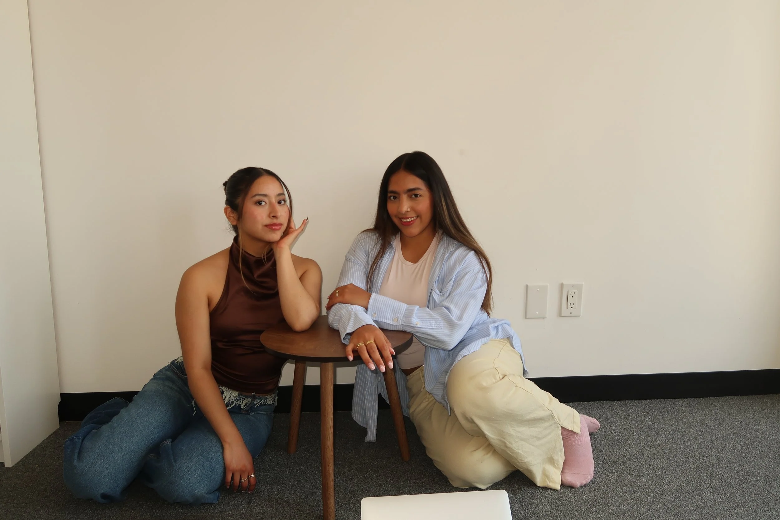 Two women sitting on the floor with their heads resting on their hands, positioned on either side of a small wooden table, smiling at the camera against a plain white wall with electrical outlets.