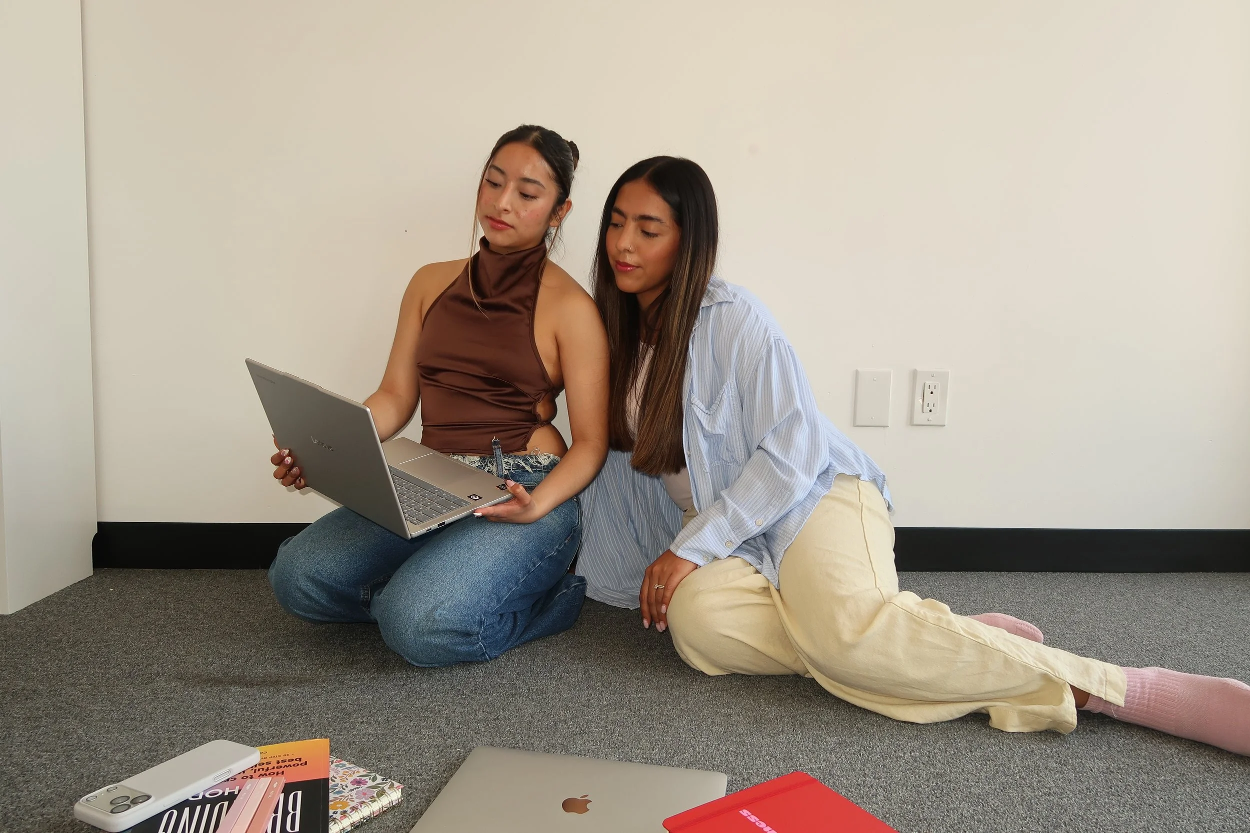 Two women are sitting on the floor against a white wall, looking at a laptop together. One woman is wearing a brown top and jeans, and the other is wearing a light blue striped shirt and beige pants. Various objects, including a phone, notebooks, and a red folder, are scattered on the floor in front of them.