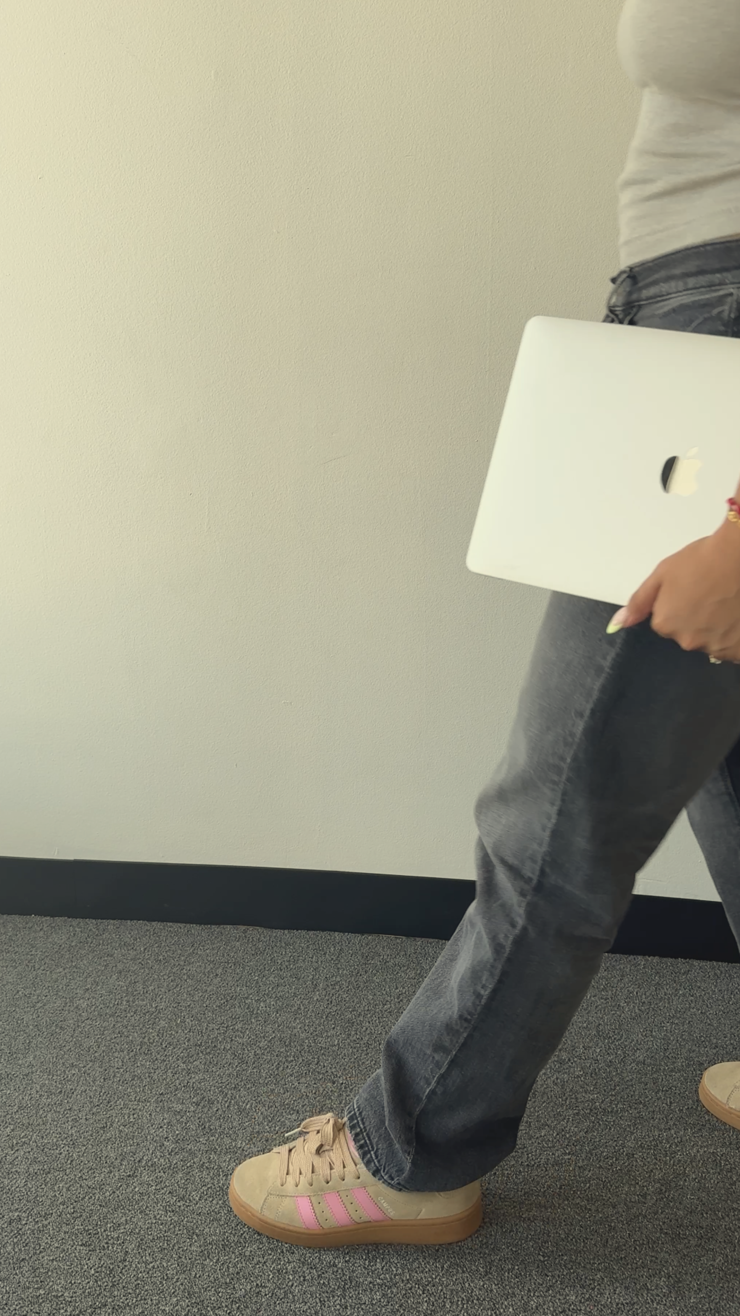 Person standing with one foot forward, wearing beige sneakers with pink stripes, gray jeans, a gray top, and holding a white laptop.