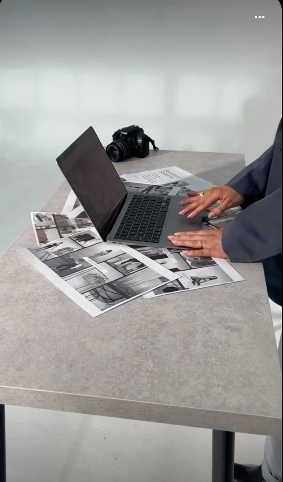 A person working on a laptop with black-and-white photographs and a camera on a table.