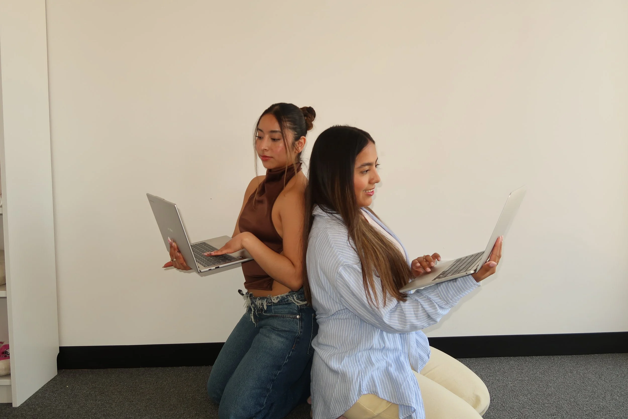 Two women sitting back-to-back, each holding and working on a laptop, against a plain beige wall.