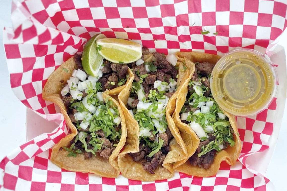 Three carne asada tacos topped with chopped onions and cilantro, lime wedges, and a small container of green salsa on red checkered paper.