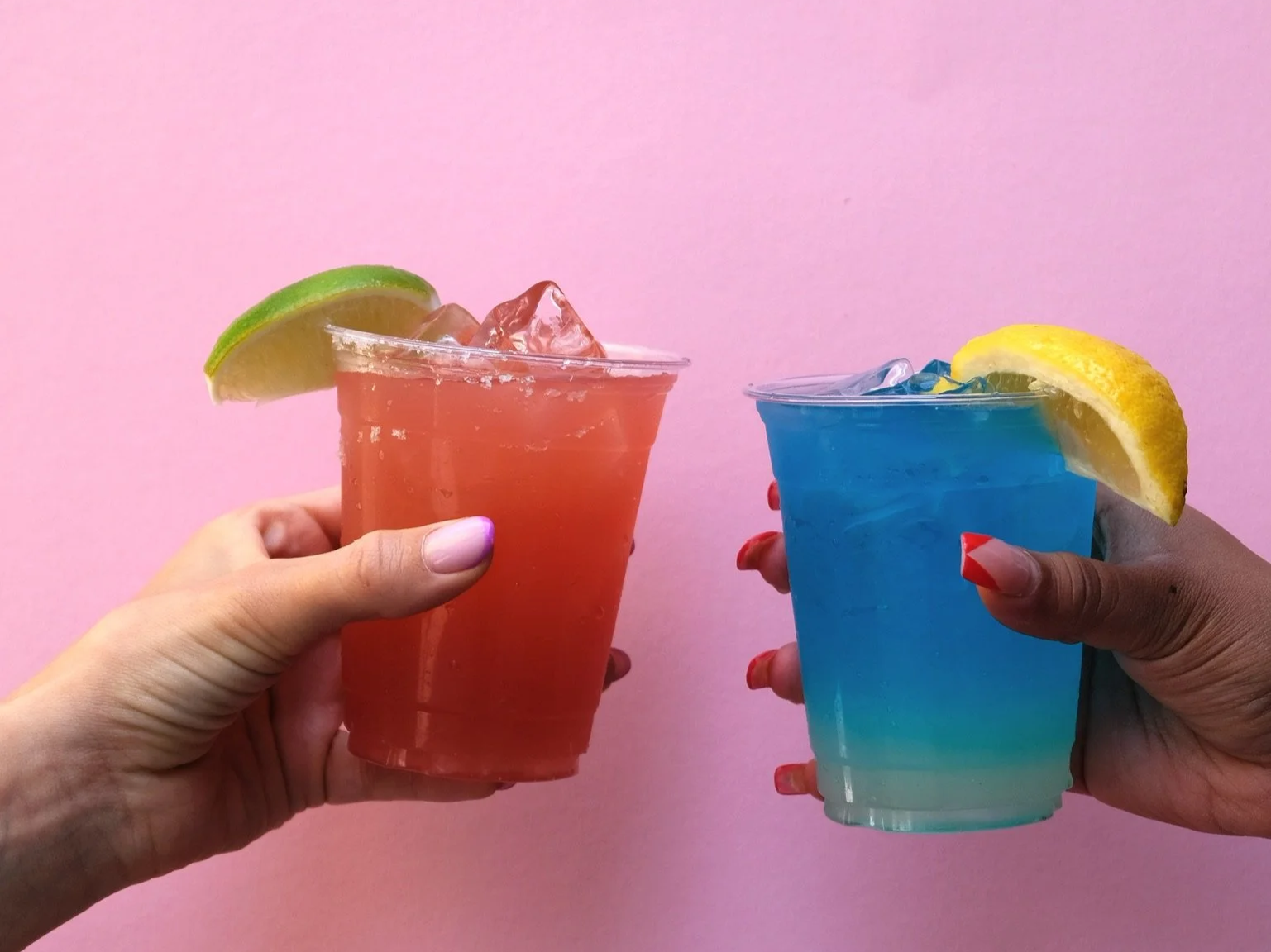Two hands holding colorful cocktails with lemon and lime wedges against a pink background.