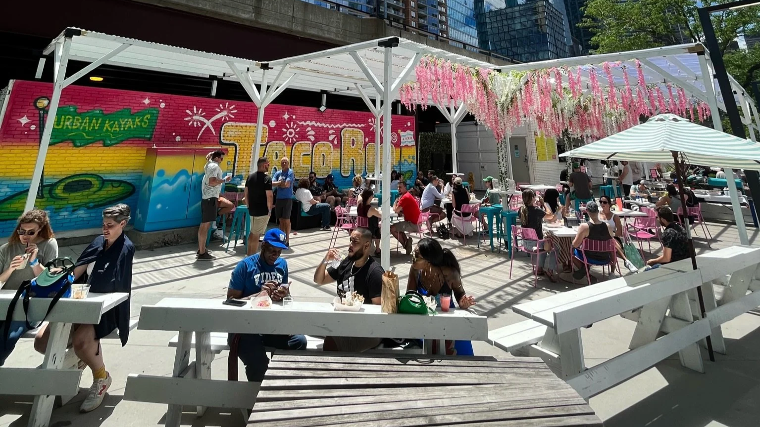 People sitting at outdoor tables with pink and blue chairs under a white pergola decorated with pink hanging flowers in an urban cafe setting
