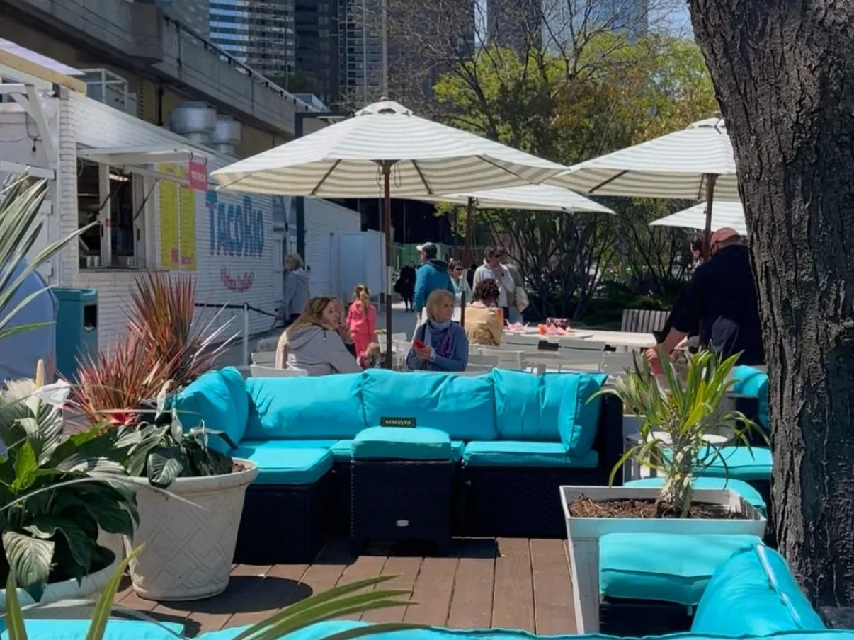 Outdoor seating area at a cafe with blue cushions, white parasols, potted plants, and people dining outside on a sunny day.