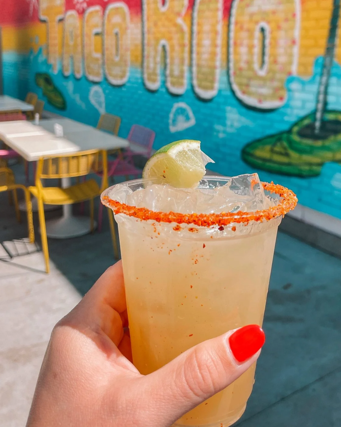 A hand with red nail polish holding a glass of light-colored beverage garnished with a lemon wedge, with a chili powder rim, in front of a colorful mural with the word 'TACO' and a taco bell mascot.