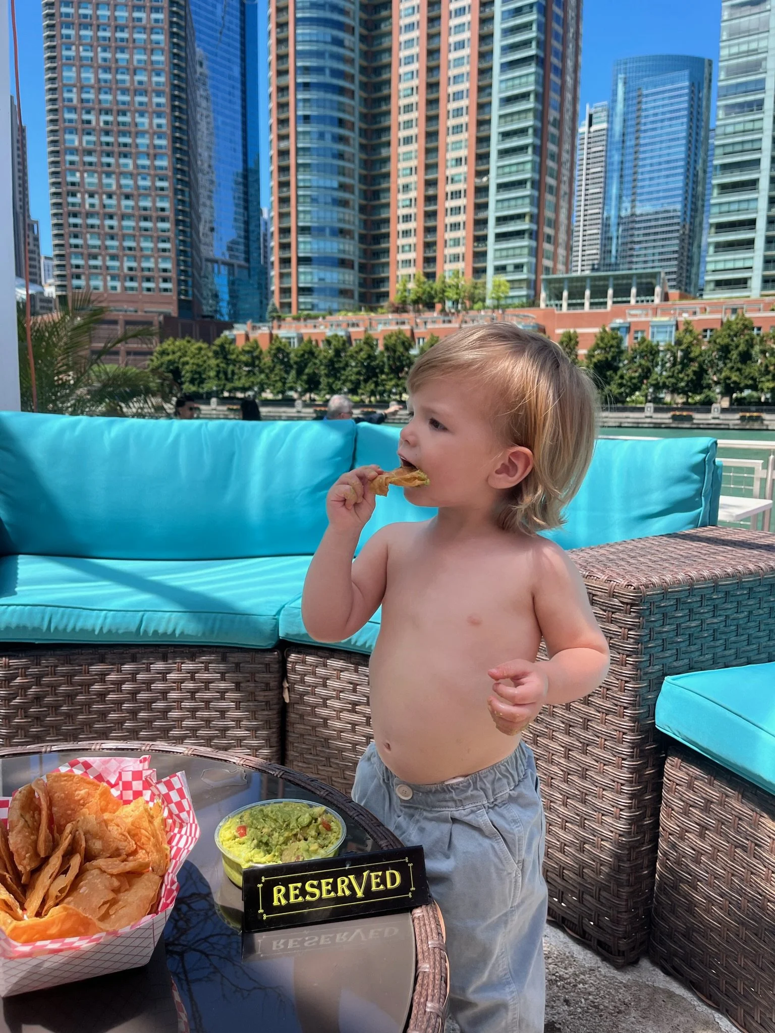 Young shirtless boy with blonde hair eating food at an outdoor restaurant with a city skyline of tall buildings in the background.