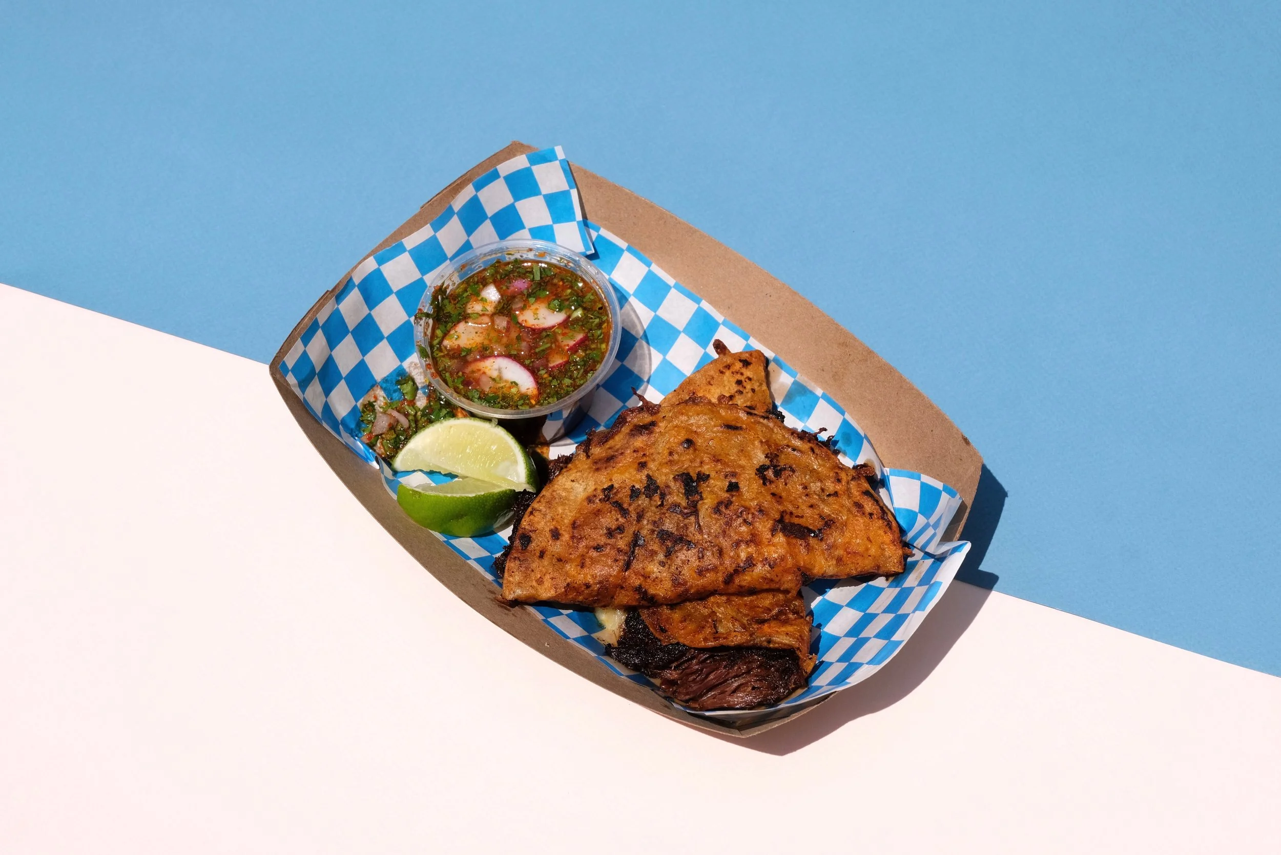 Mexican-style grilled meat with lime wedges and salsa in a paper basket with a blue and white checkered liner on a blue and white background.
