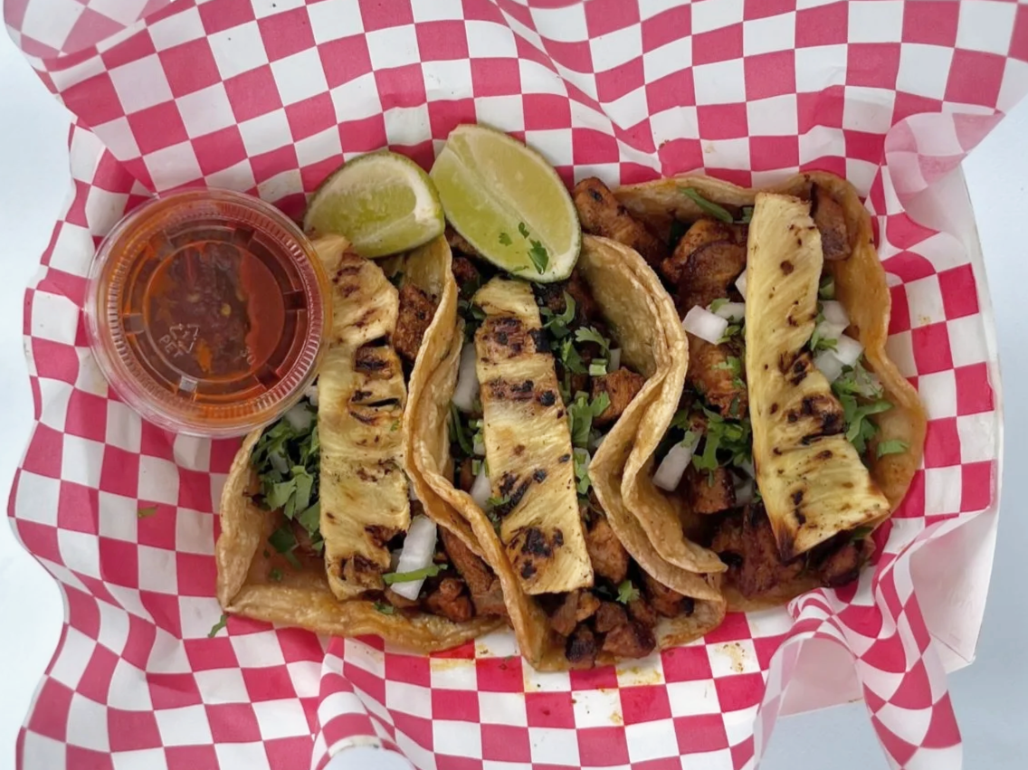 Three grilled steak tacos with grilled pineapple slices, chopped onions, cilantro, and lime wedges, served with a side of salsa in a small cup.
