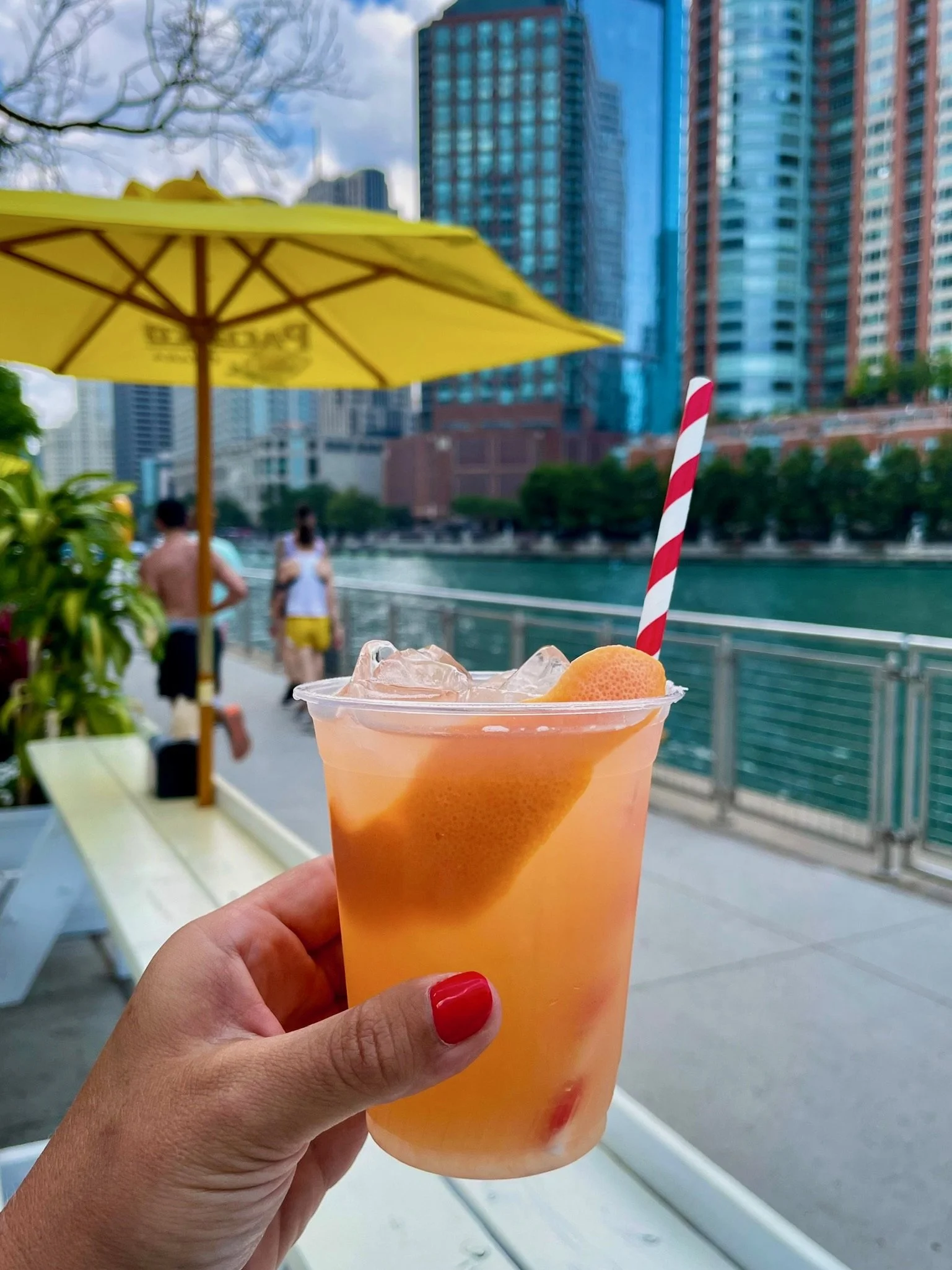 A hand holding an orange-colored cocktail with ice, a slice of citrus fruit, and a red-and-white striped straw, against an urban riverside backdrop with buildings, trees, and a yellow umbrella.